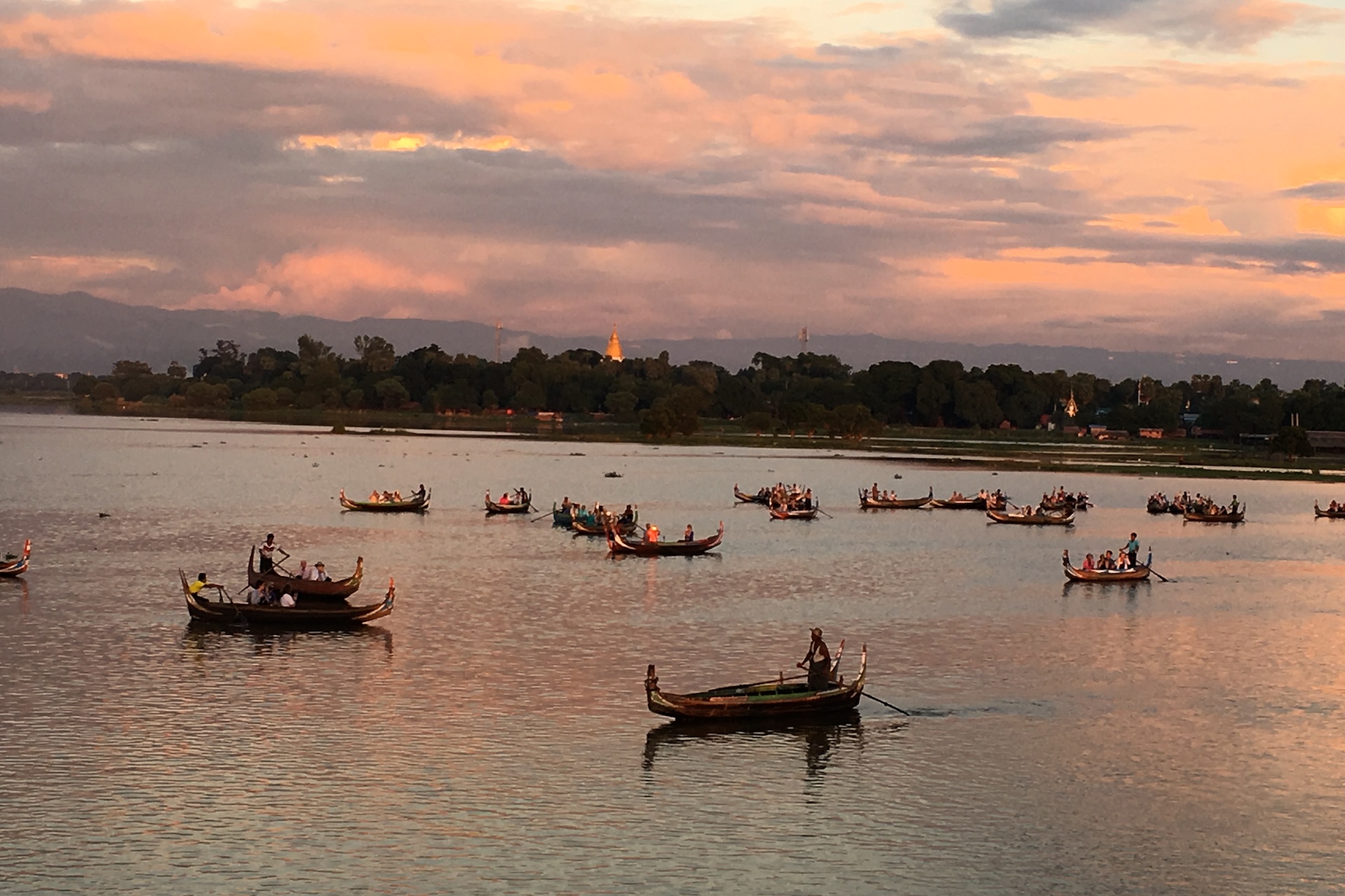 Boats carrying people drift across Amarapura - Taungthaman Lake at sunset near U Bein Bridge in Mandalay, Myanmar, with a golden pagoda visible in the distance.