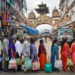 Nepal NRs 100 Customs Crackdown: People standing in long queue with bags at India–Nepal border checkpoint as trucks pass through decorated market street amid strict NRs 100 customs enforcement