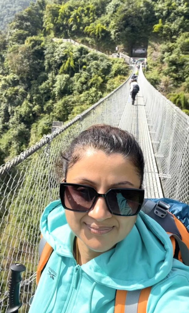 Trekker taking a selfie while crossing the Jhinu Danda steel wire suspension bridge on Day 1 of the ABC Trek in Nepal, surrounded by lush green Himalayan forest.
