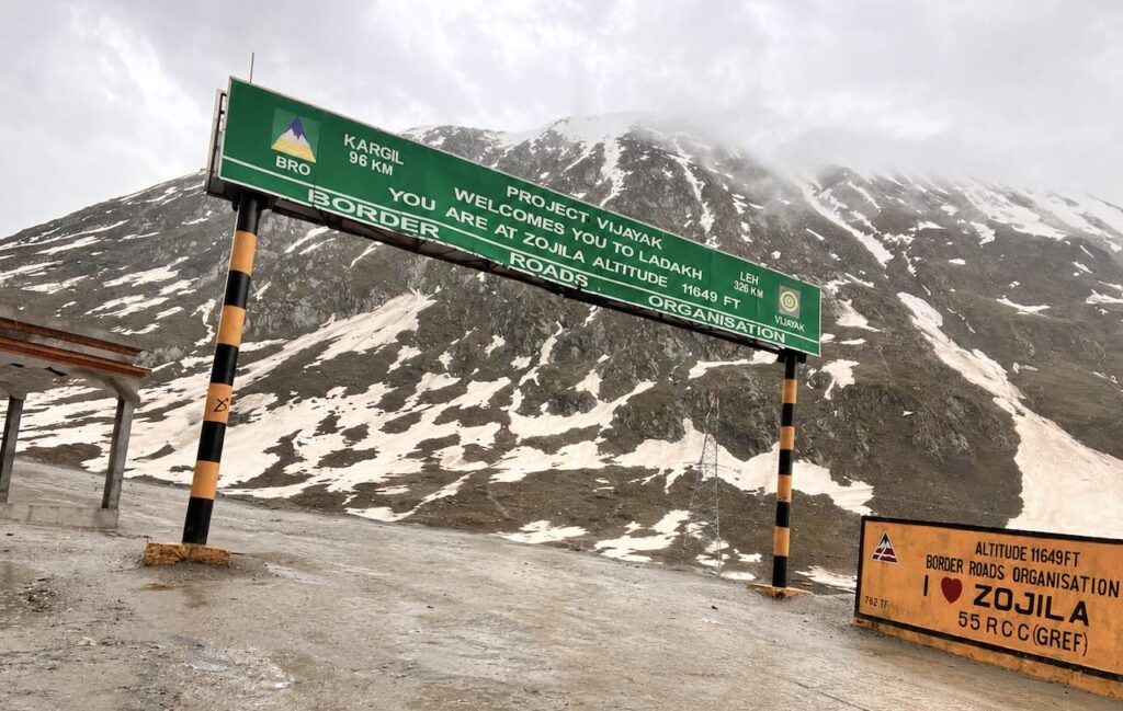 Green BRO welcome sign at Zojila Pass standing against rugged snow-covered Himalayan mountains under cloudy skies.